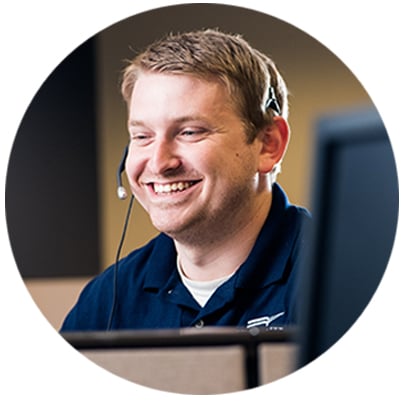An emergency dispatcher in a blue shirt smiles while sitting at a computer talking on a bluetooth headset